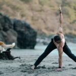 Doga: homem fazendo postura de yoga na praia com dois cães brincando ao fundo